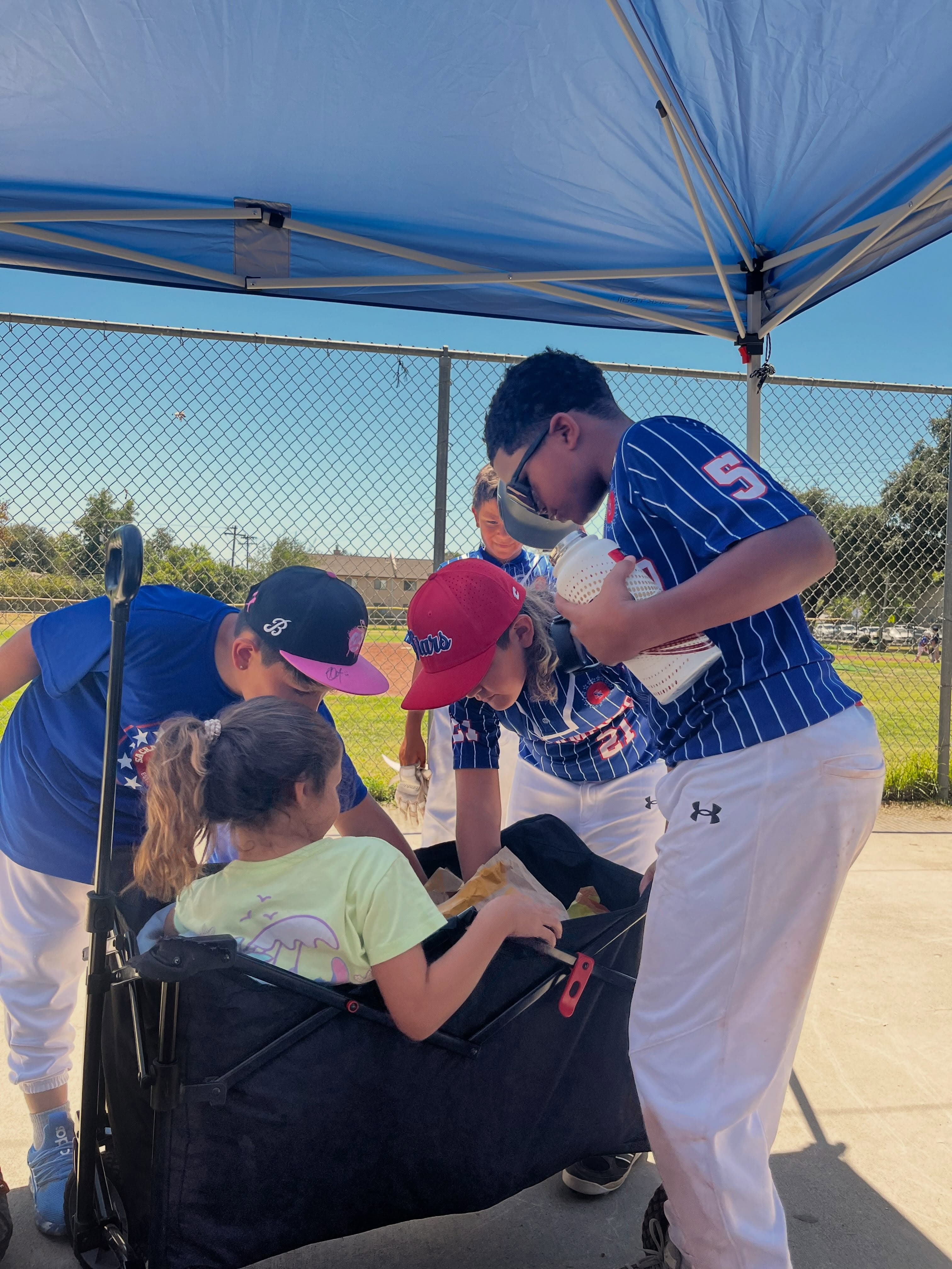 Players gather under a tent with a younger sibling in a wagon after a game.